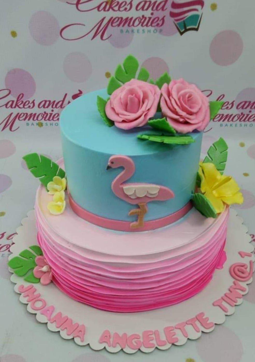 Two-tier tropical flamingo cake with pink ruffles, blue top tier, and edible pink roses and tropical leaves.