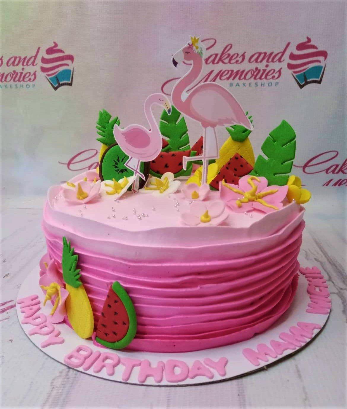 Pink flamingo tropical birthday cake with watermelon, pineapple, and kiwi fondant decorations and pink flowers.