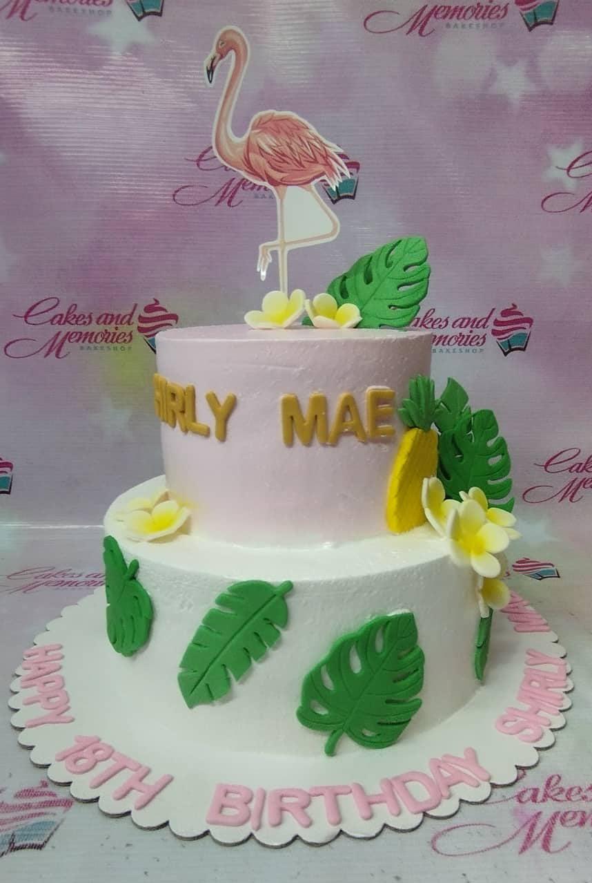 Two-tier tropical flamingo birthday cake with pink and white icing, fondant monstera leaves, and yellow frangipani flowers.