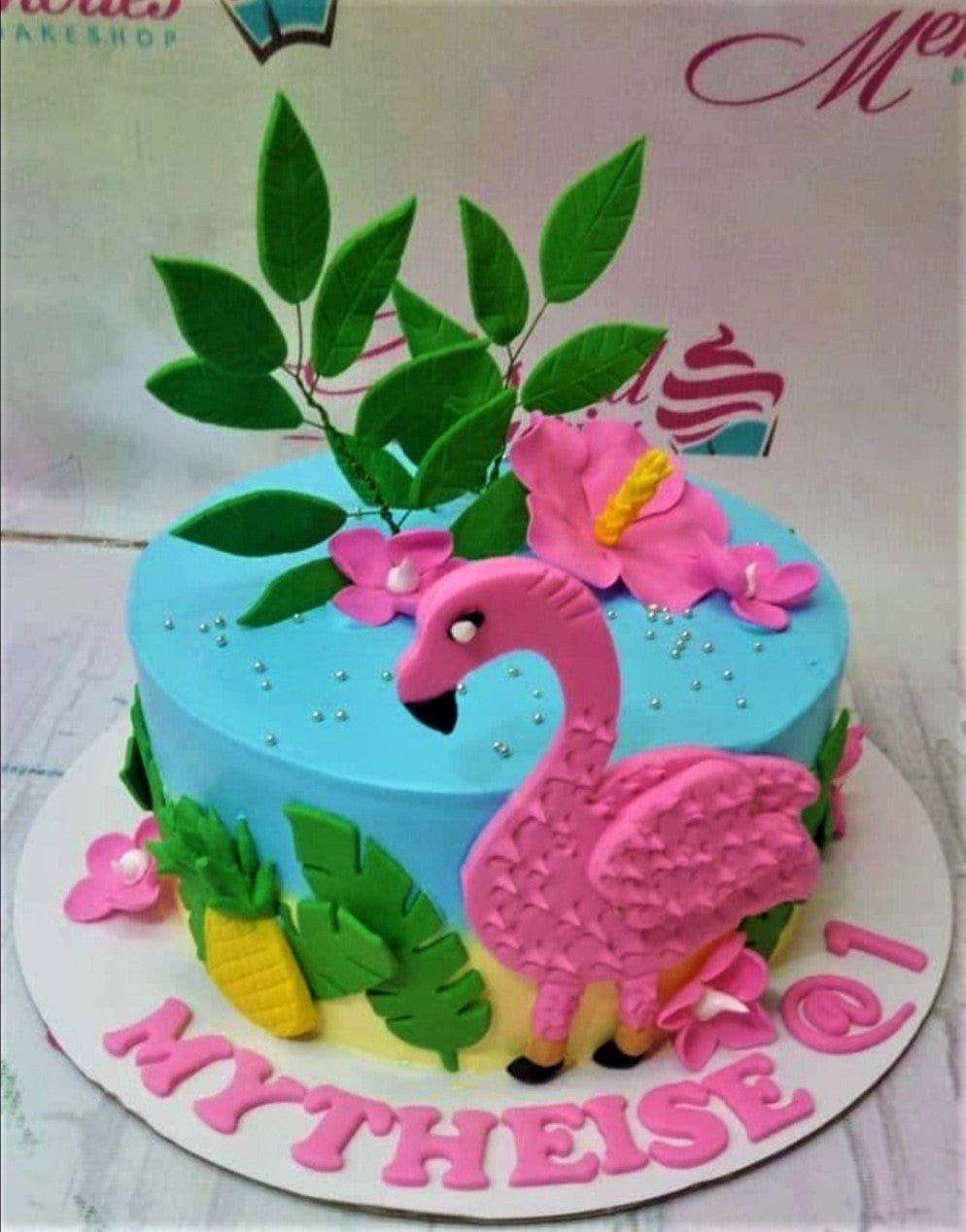 Tropical flamingo birthday cake with pink fondant flamingo, green leaves, and pink hibiscus flowers on light blue icing.