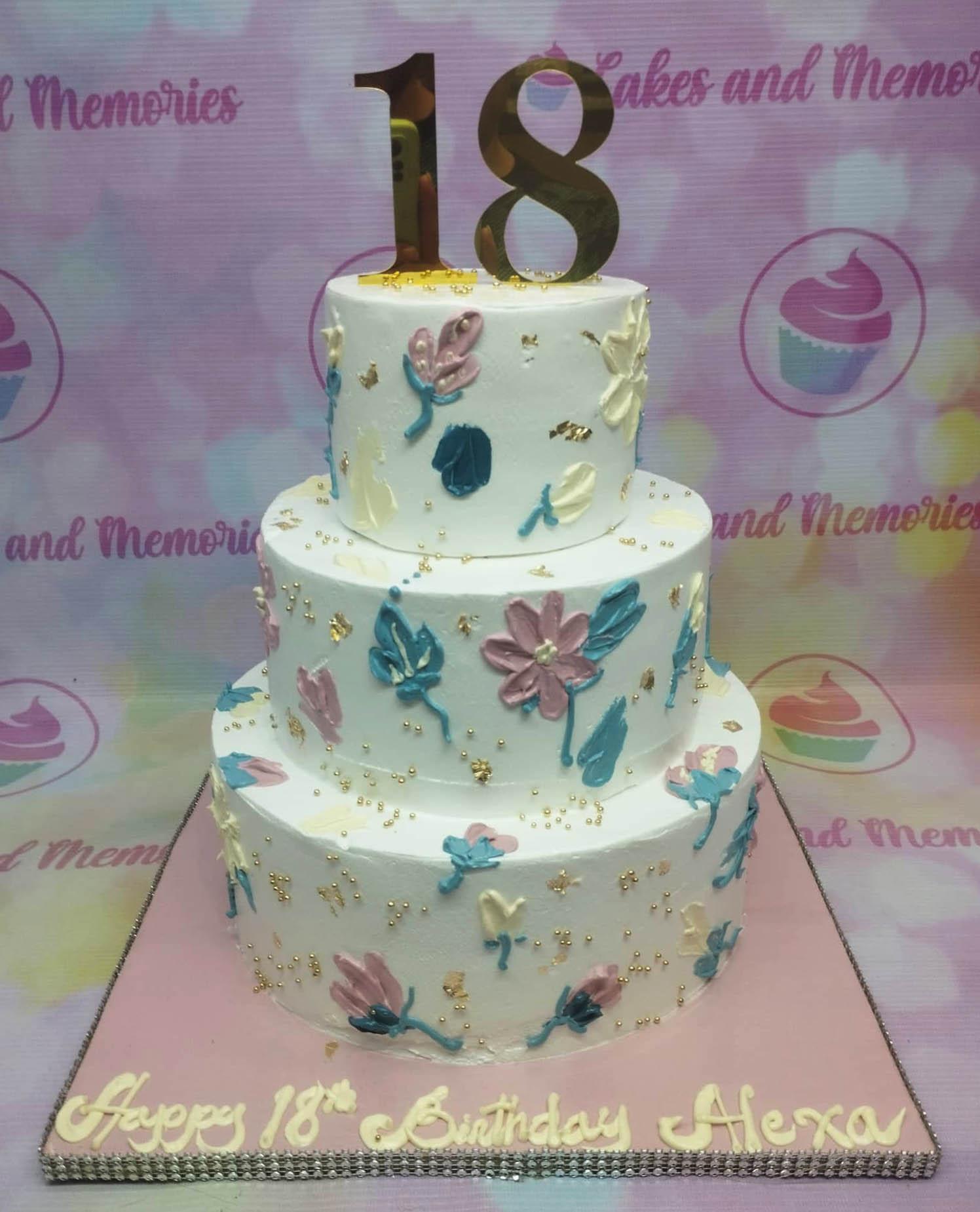 Elegant 3-tier white floral cake with palette knife icing flowers in pink, blue, and yellow, topped with a gold number 18.