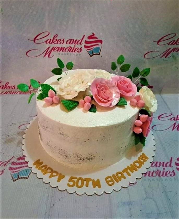 Semi-naked white birthday cake decorated with pink and white gumpaste roses, green leaves, and pink berries with a gold message on the base.