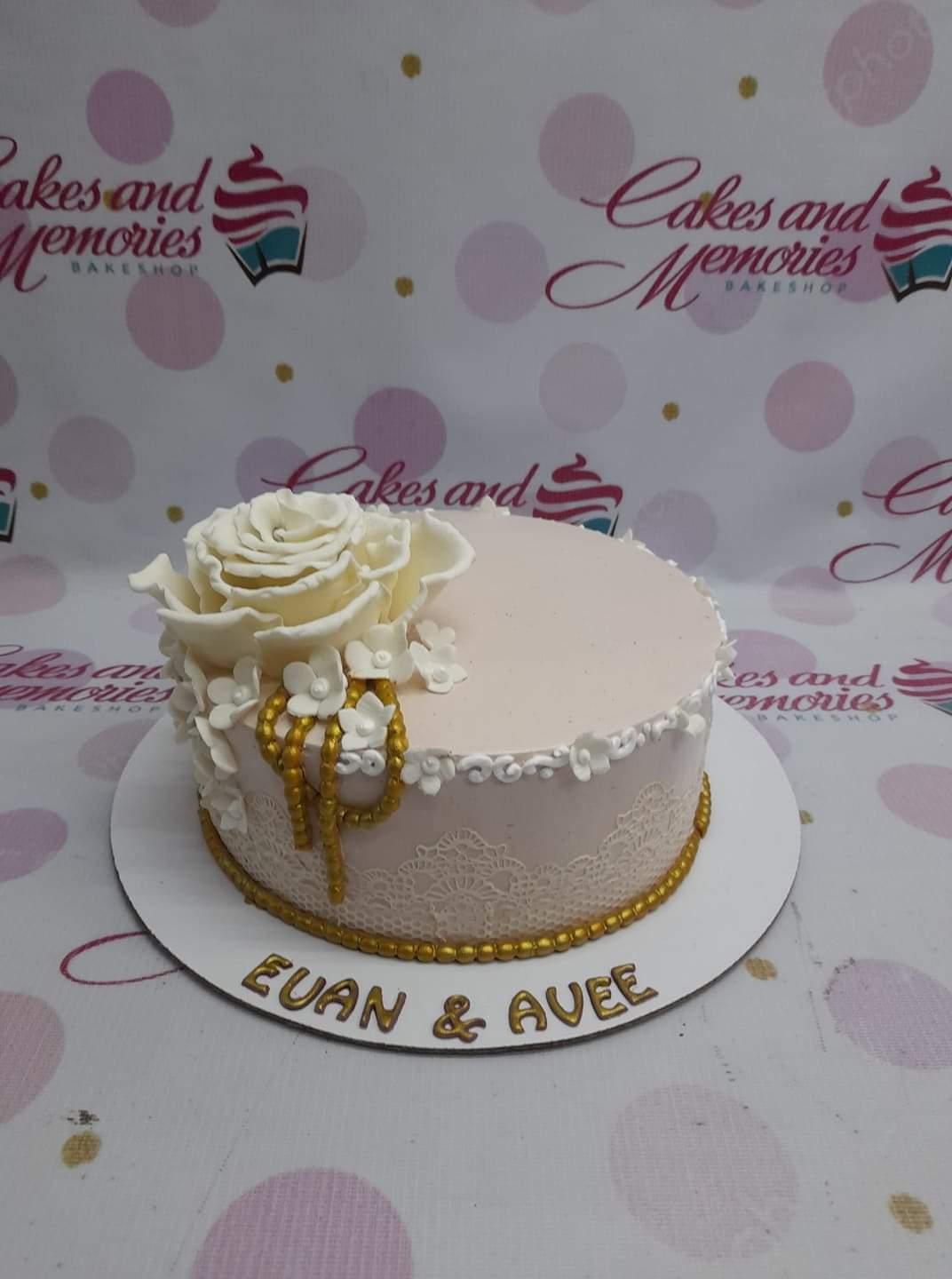Elegant light pink single tier cake with a large white gumpaste rose, small white flowers, gold beads, and white lace icing details.