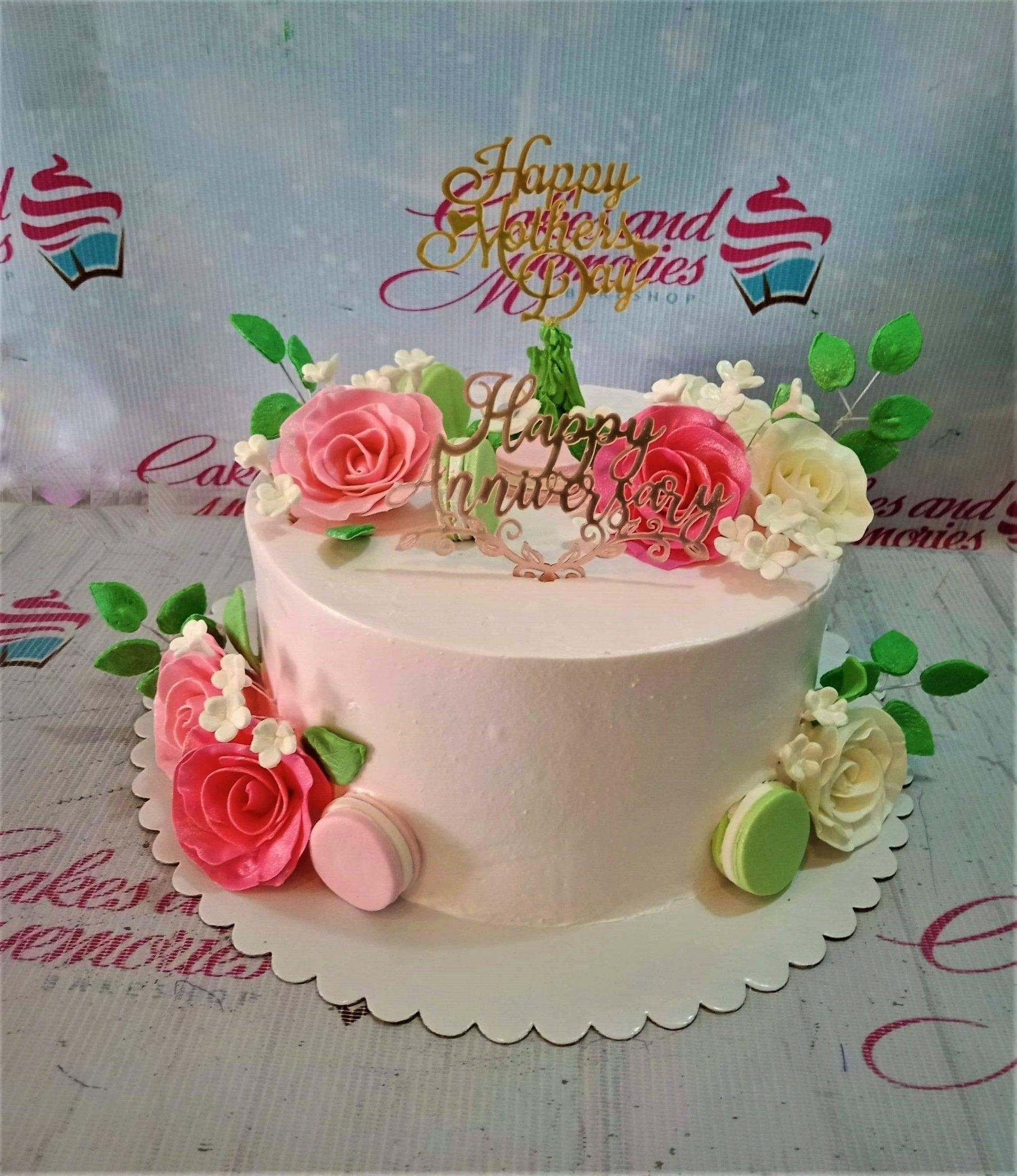 White soft-icing anniversary cake decorated with pink and white gumpaste roses, green leaves, macarons, and gold toppers.