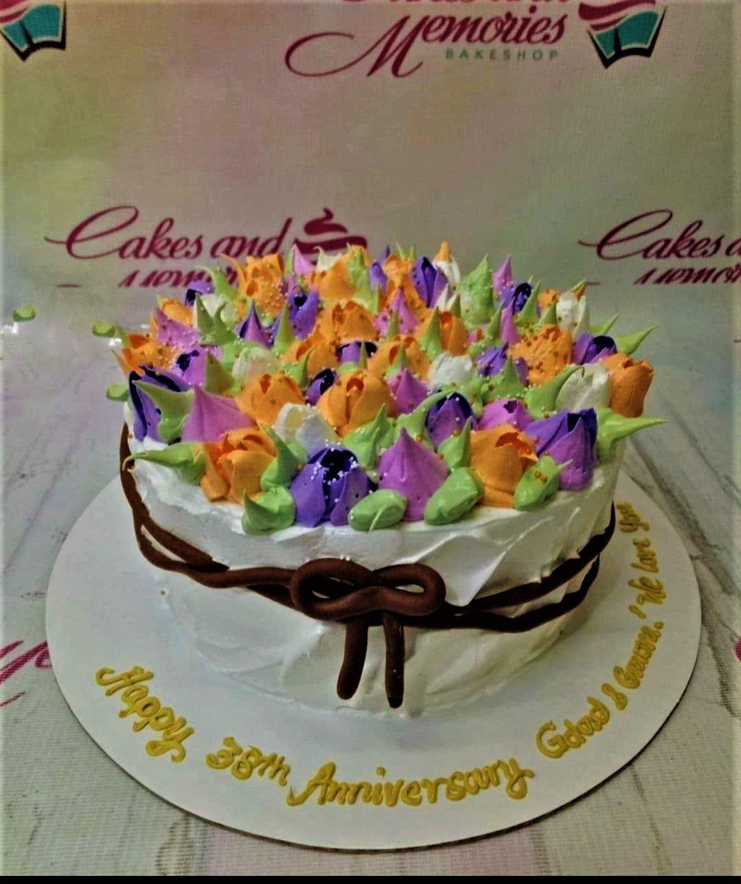 White soft-icing cake topped with a dense garden of purple, orange, and white icing flowers with green leaves and a brown icing bow.