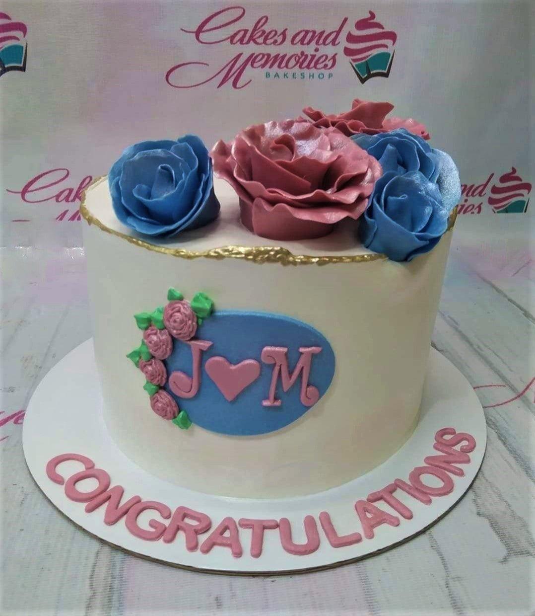 White single tier cake with large blue and pink gumpaste roses, gold rim, and a blue oval plaque with pink initials J and M.