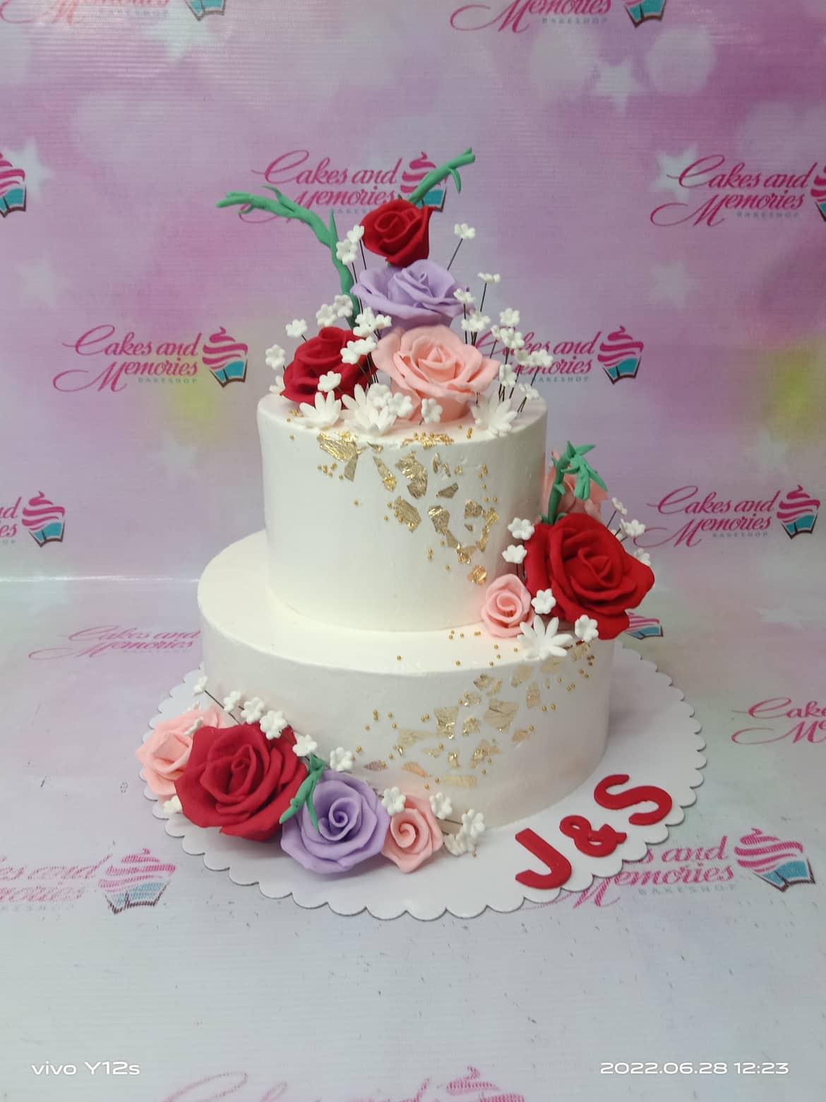 Elegant 2-tier white floral cake with red, purple, and pink gumpaste roses, gold leaf accents, and red gumpaste initials J&S.