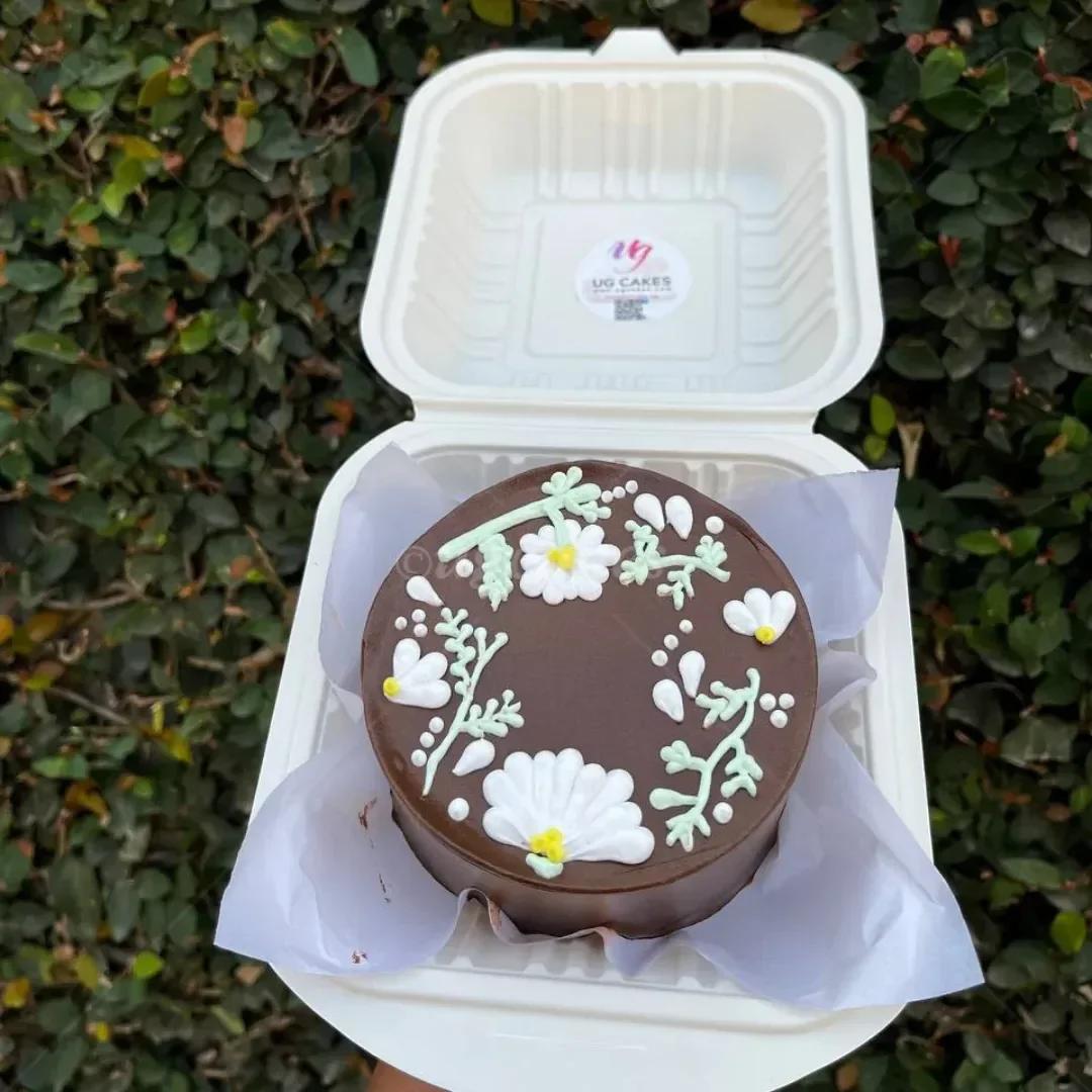 A chocolate bento cake with white and yellow piped icing flowers and light green stems in a white takeout container.