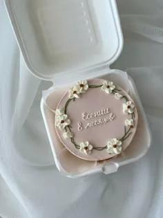 Light pink bento cake with a white piped floral wreath and green stems, featuring a white icing message in the center.