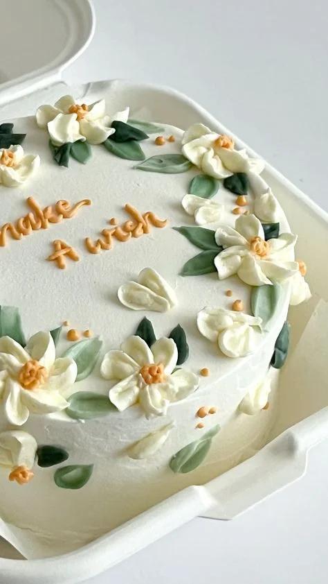 White bento cake with piped ivory icing flowers, green leaves, and peach centers, featuring the message 'Make A Wish' in peach icing.