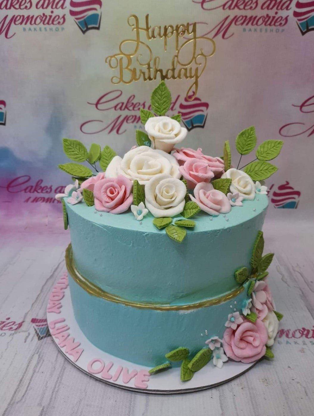 Two-tier light blue floral cake with pink and white gumpaste roses, green leaves, and a gold Happy Birthday topper.