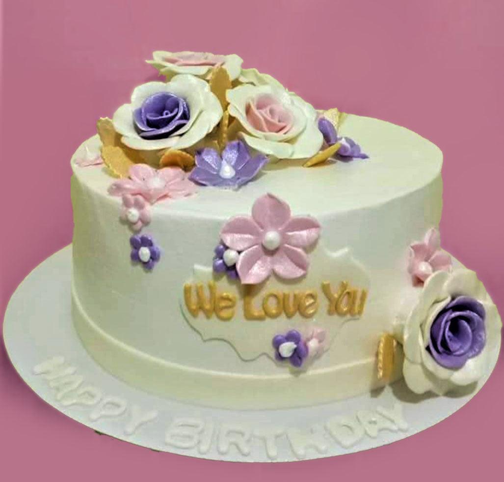 White round cake decorated with purple and pink gumpaste roses and flowers with gold leaves and a We Love You message.
