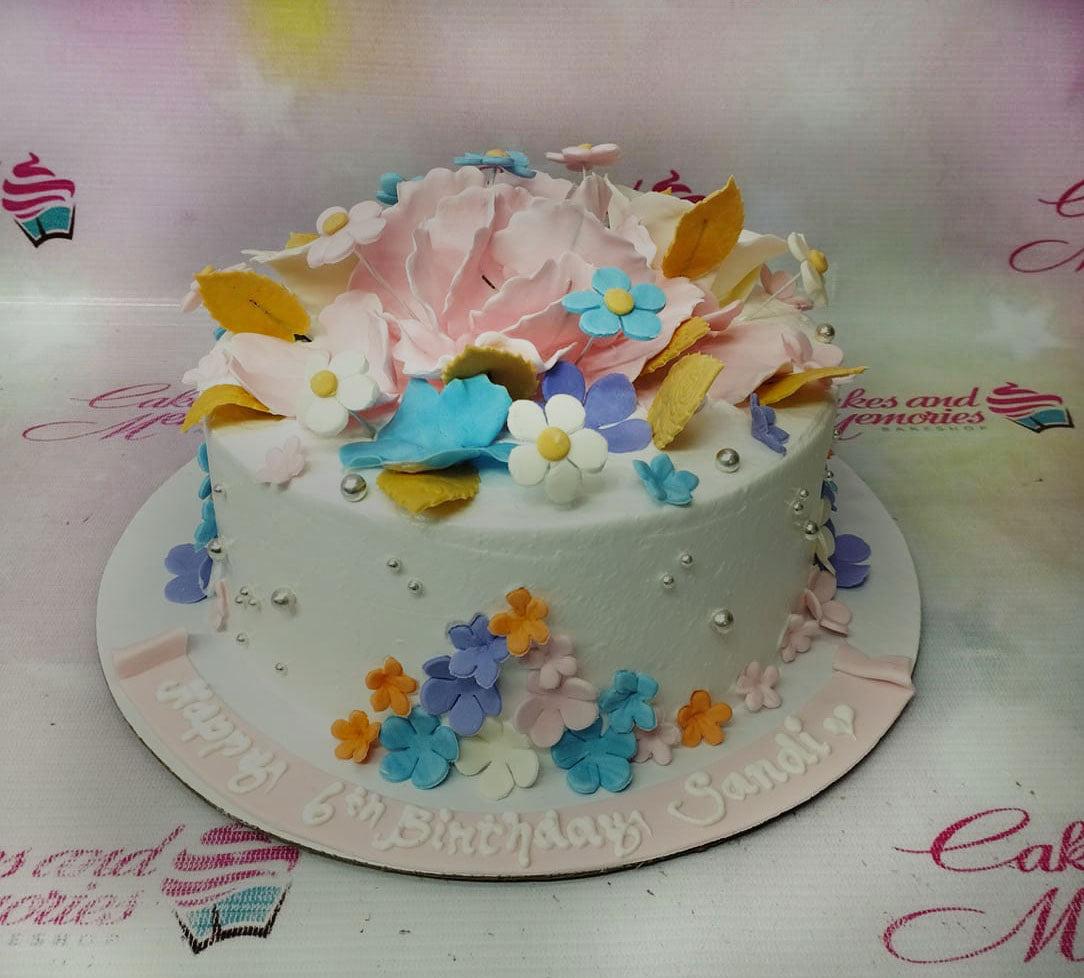 White round birthday cake decorated with pink, blue, and purple gumpaste flowers and silver dragees with a pink message ribbon.