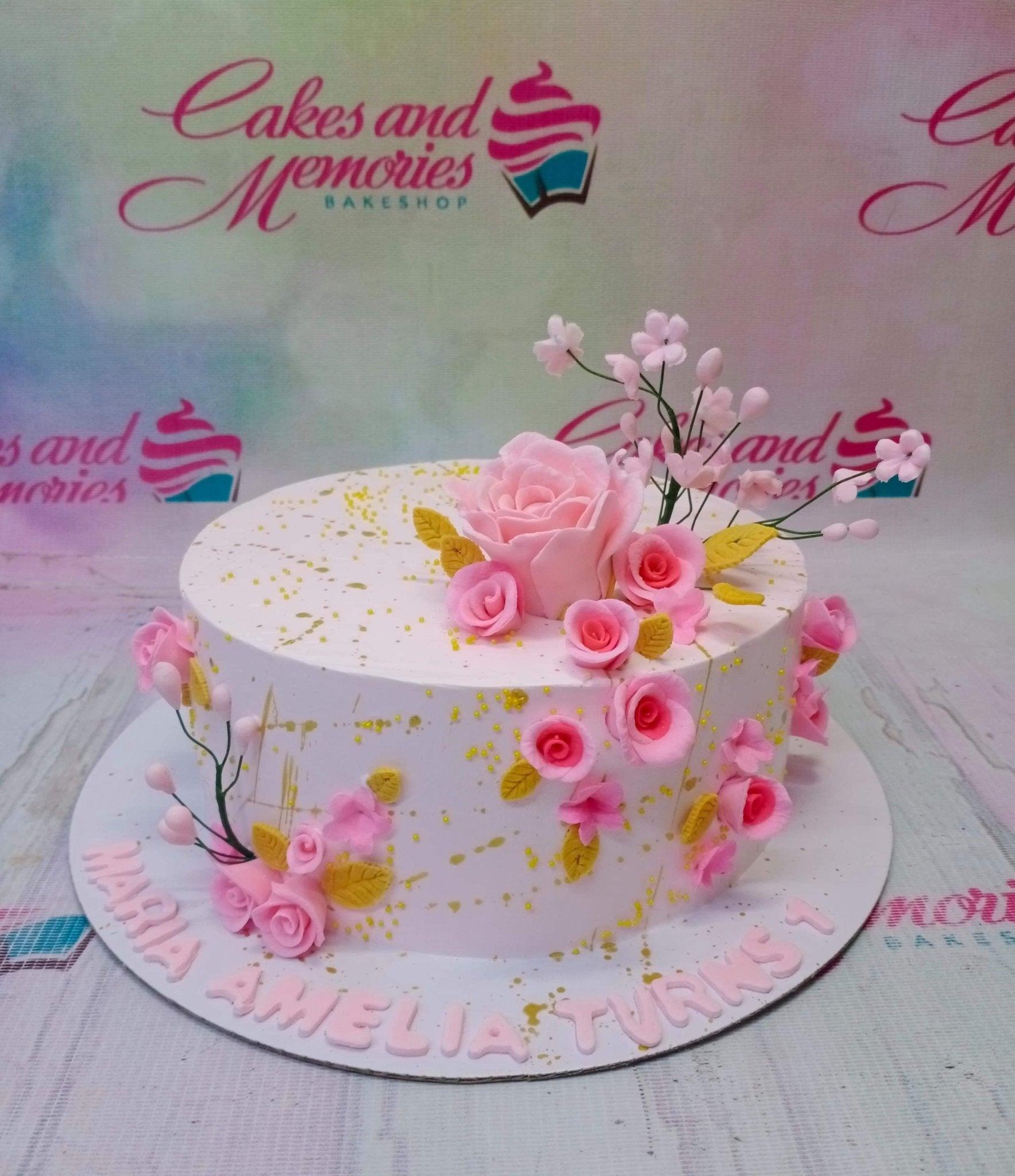 White 1-tier floral birthday cake with pink fondant roses, gold splatters, and pink gumpaste letters on the base board.