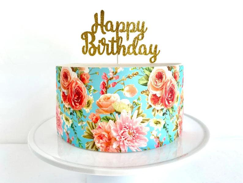 Single tier light blue cake with a vibrant floral print wrap and a gold glitter Happy Birthday topper on a white cake stand.