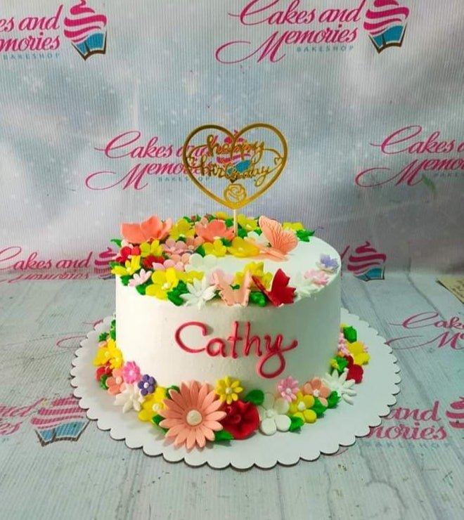 White round cake decorated with colorful gumpaste flowers and butterflies with a gold heart-shaped Happy Birthday topper.