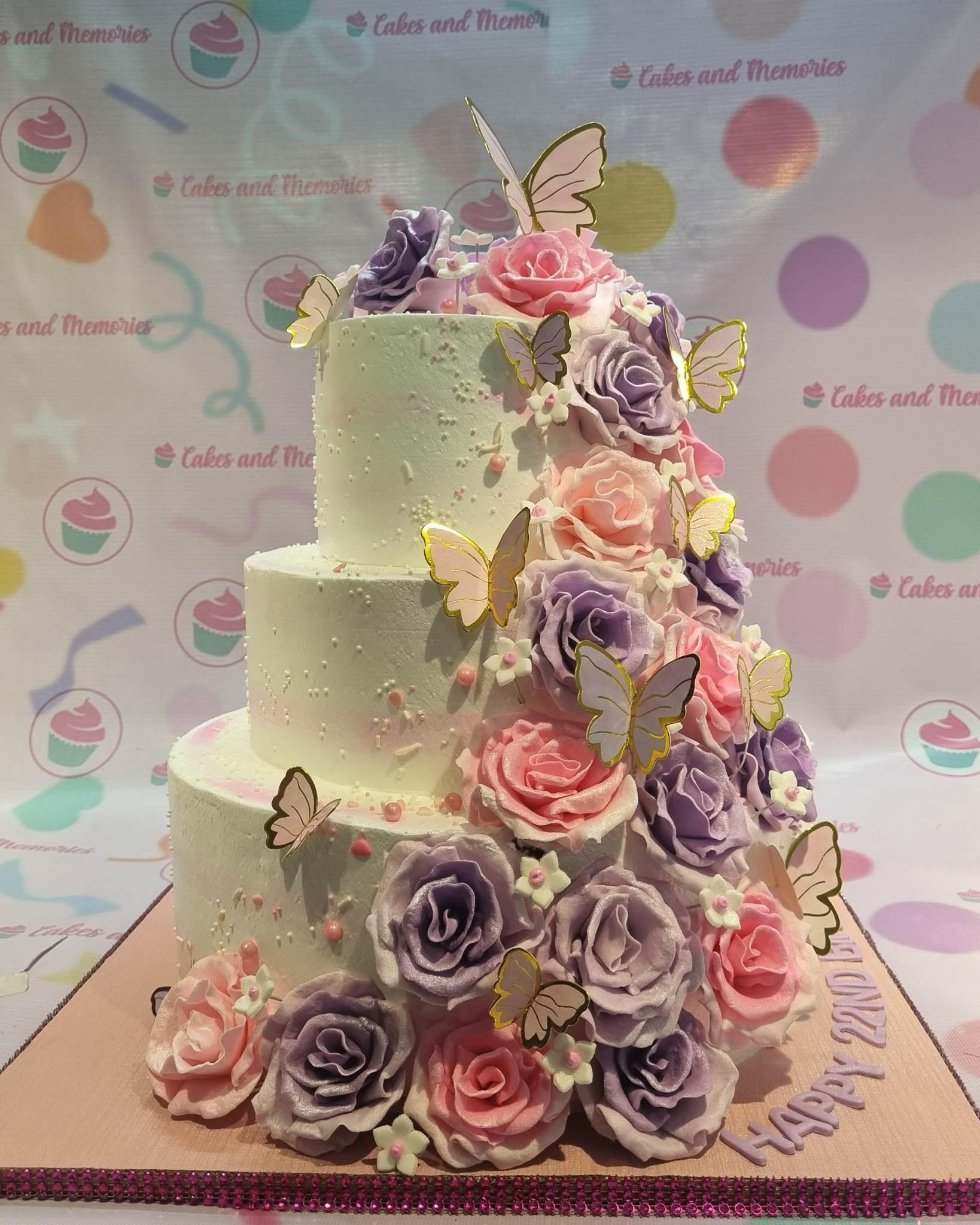 Elegant three-tier white cake adorned with a cascade of pink and purple gumpaste roses and gold-edged butterfly toppers.