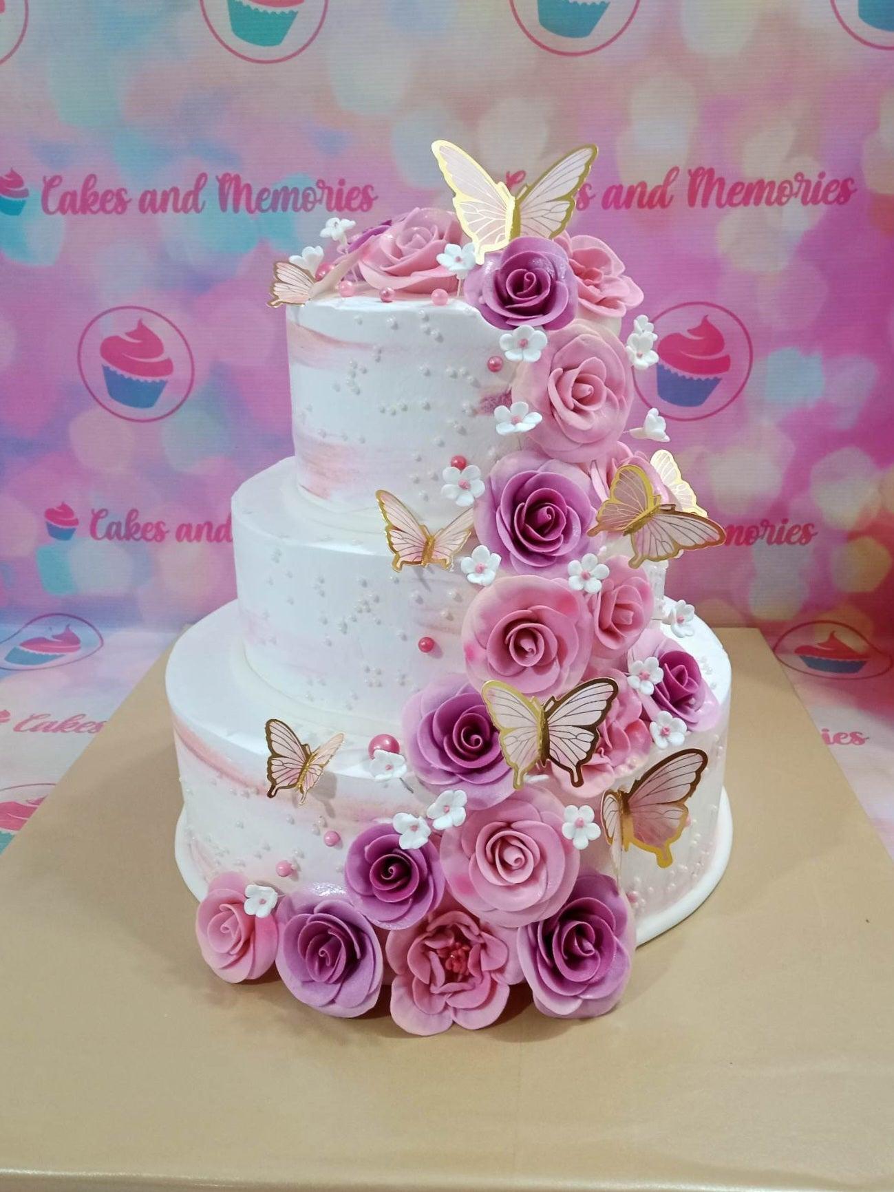 Elegant three-tier white cake with pink and purple gumpaste roses, gold butterflies, and white icing rosettes cascading down the side.