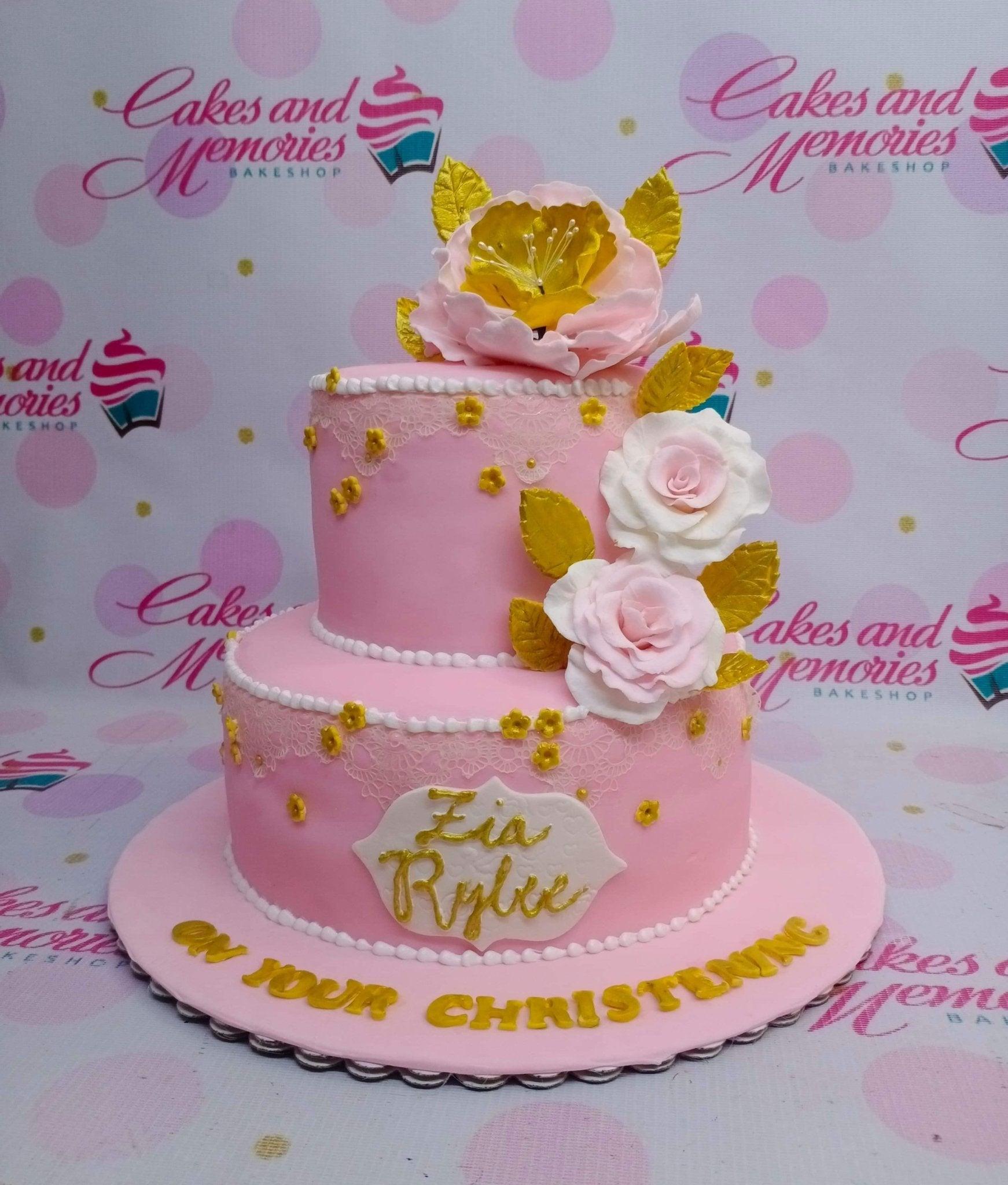 Pink 2-tier christening cake with large pink and white sugar flowers, gold leaves, and yellow floral accents.
