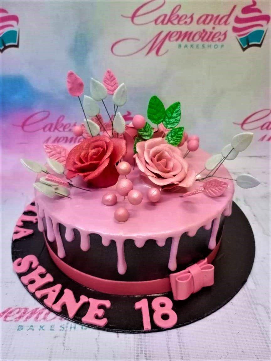 Pink and black drip cake with edible gumpaste roses and pink gumpaste letters on a black base board.