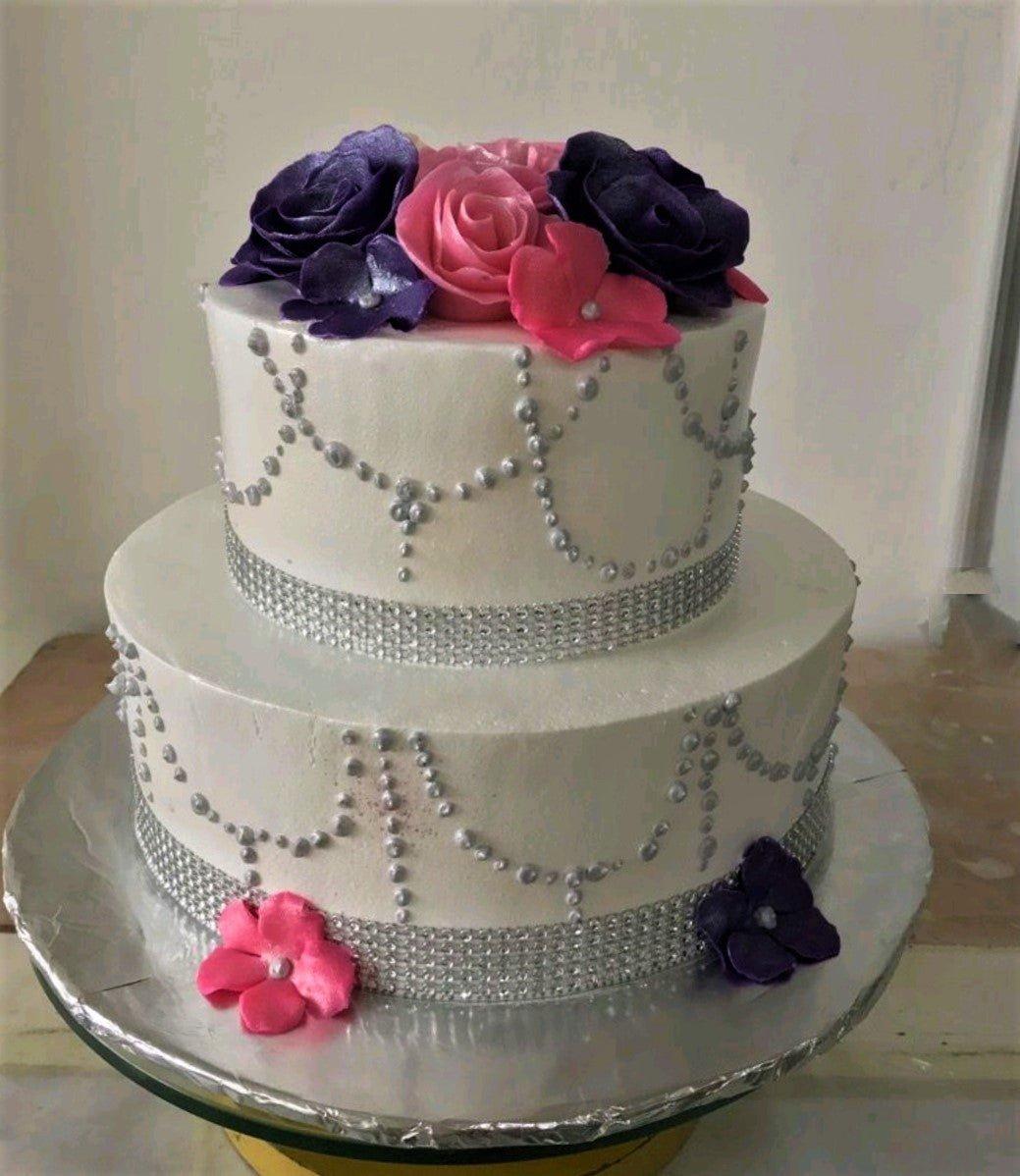Elegant 2-tier white cake with purple and pink gumpaste roses, silver bead swags, and rhinestone ribbon borders.