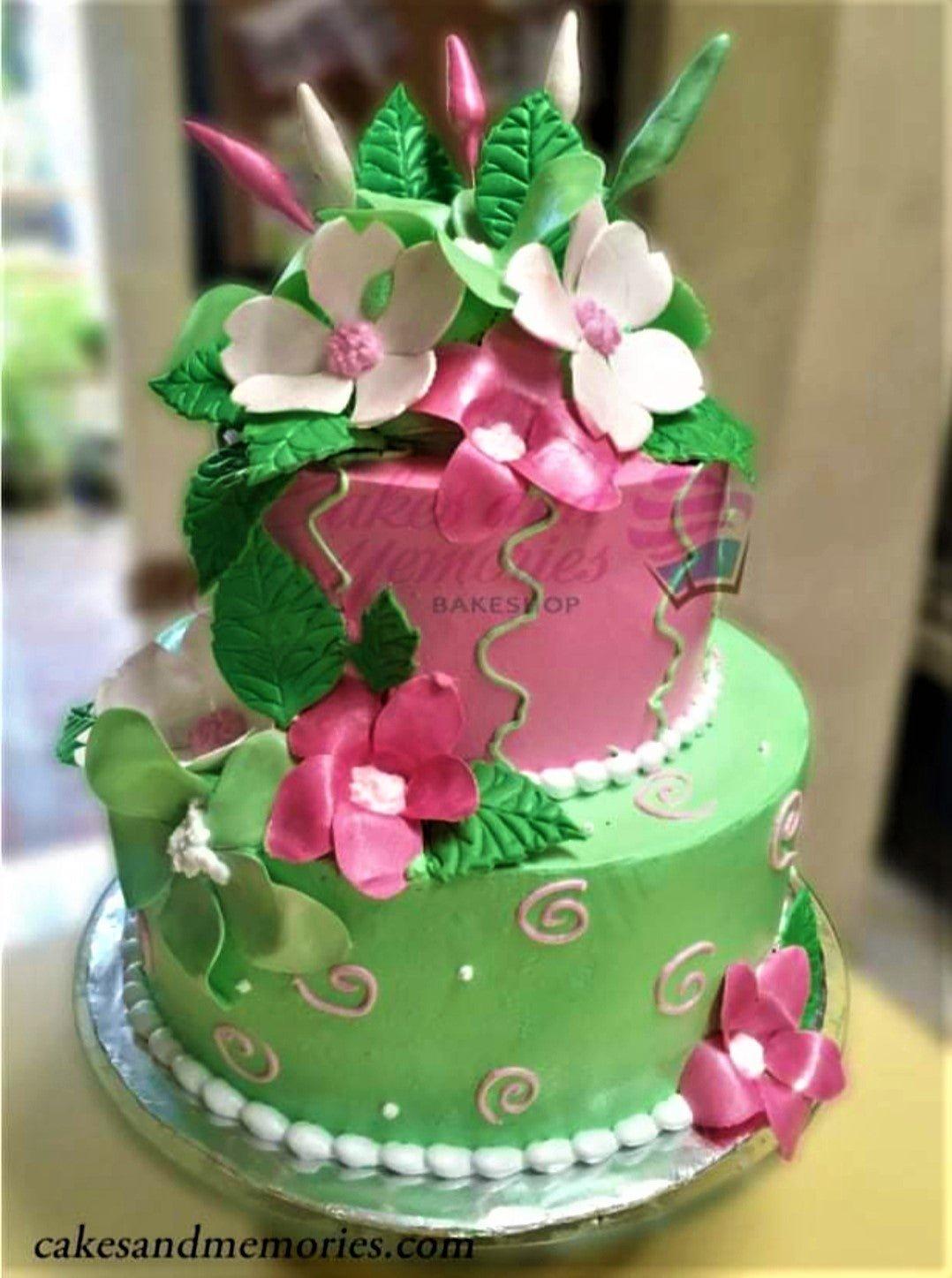 Two-tier floral cake with pink and green icing, featuring large pink and white gumpaste flowers and green leaves.