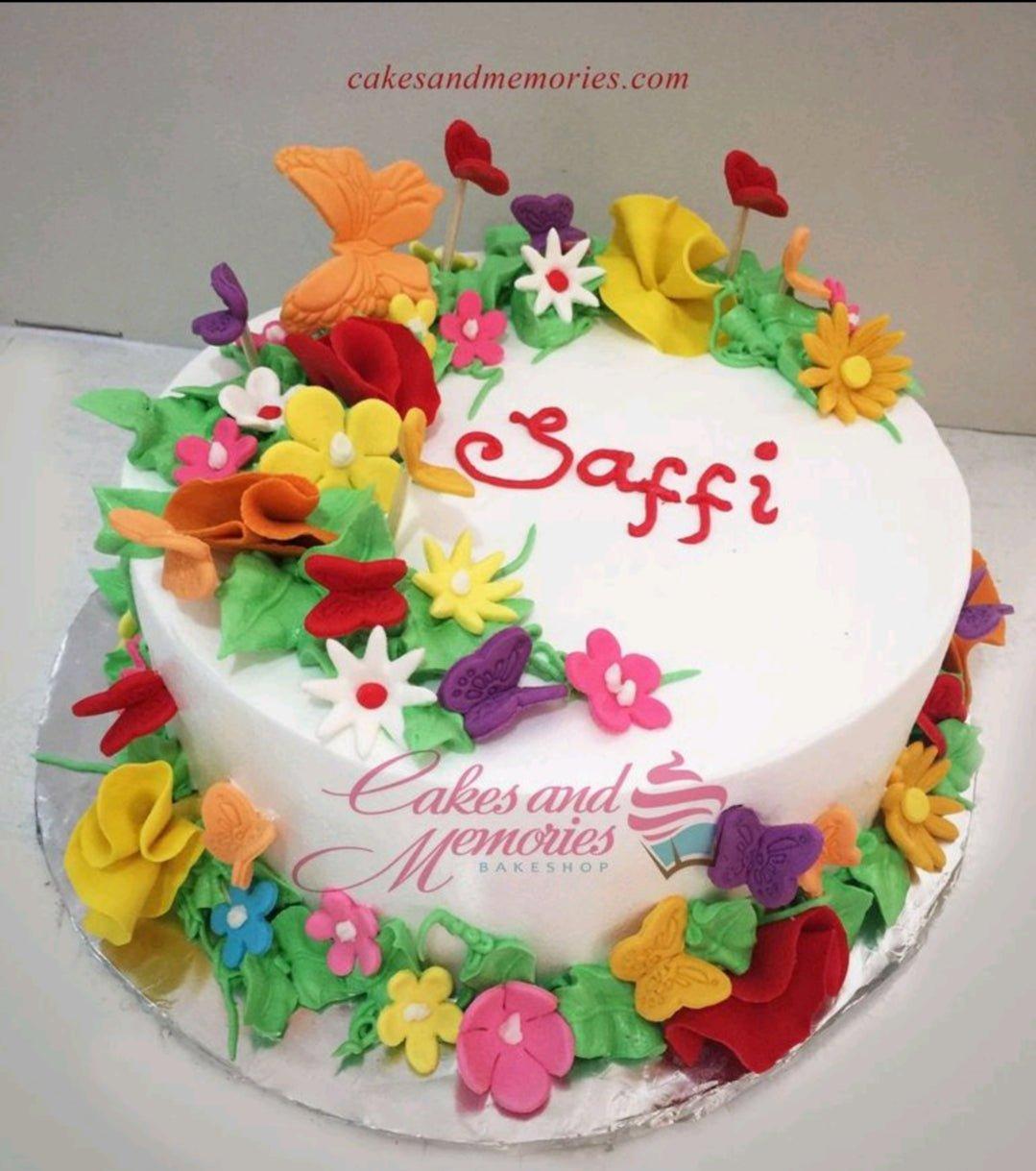 White round cake decorated with colorful gumpaste flowers, butterflies, and green leaves with red icing text Saffi.