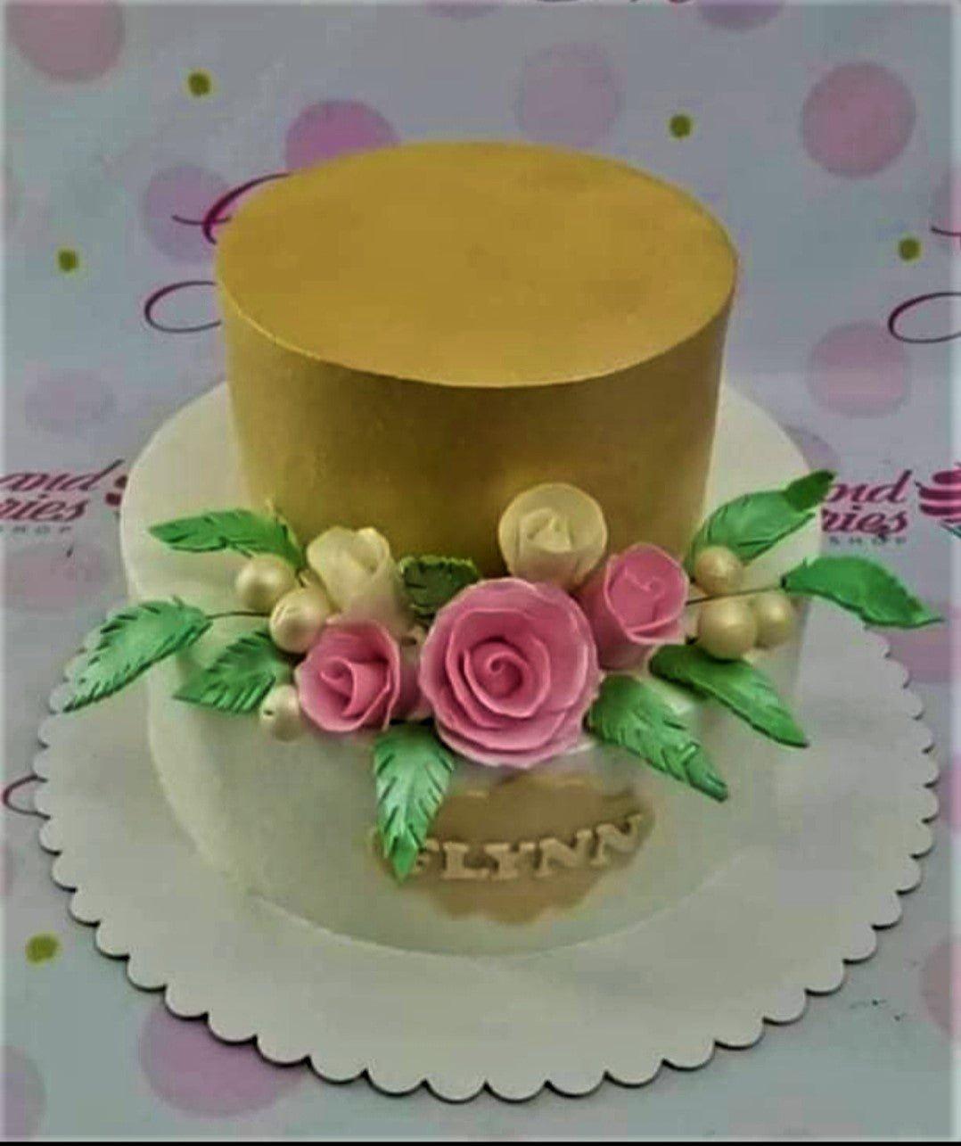 Elegant two-tier cake with a gold top tier and white bottom tier decorated with pink and cream roses and green leaves.