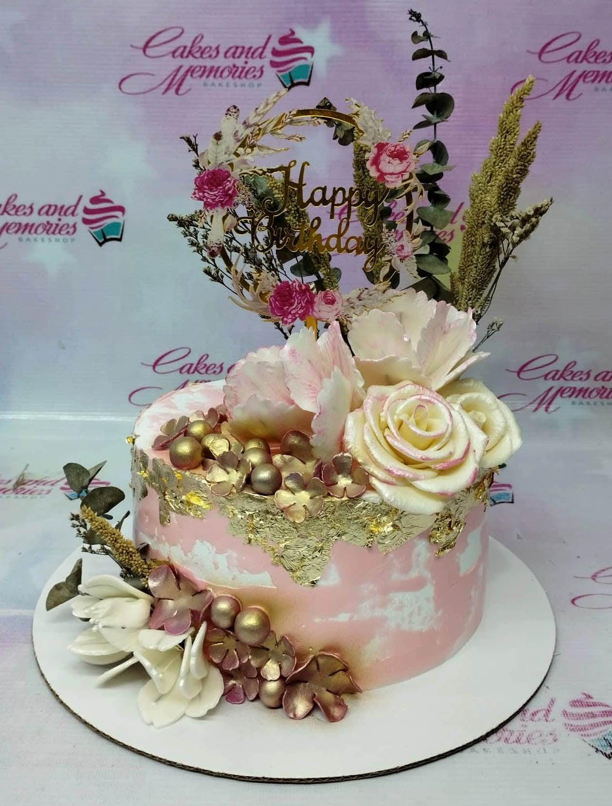 Pink and white marbled cake decorated with large white and pink edible flowers, gold leaf, and a gold Happy Birthday topper.