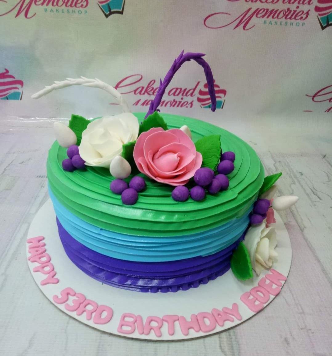A single-tier birthday cake with horizontal gradient icing in green, blue, and purple, decorated with pink and white fondant roses and purple grapes.