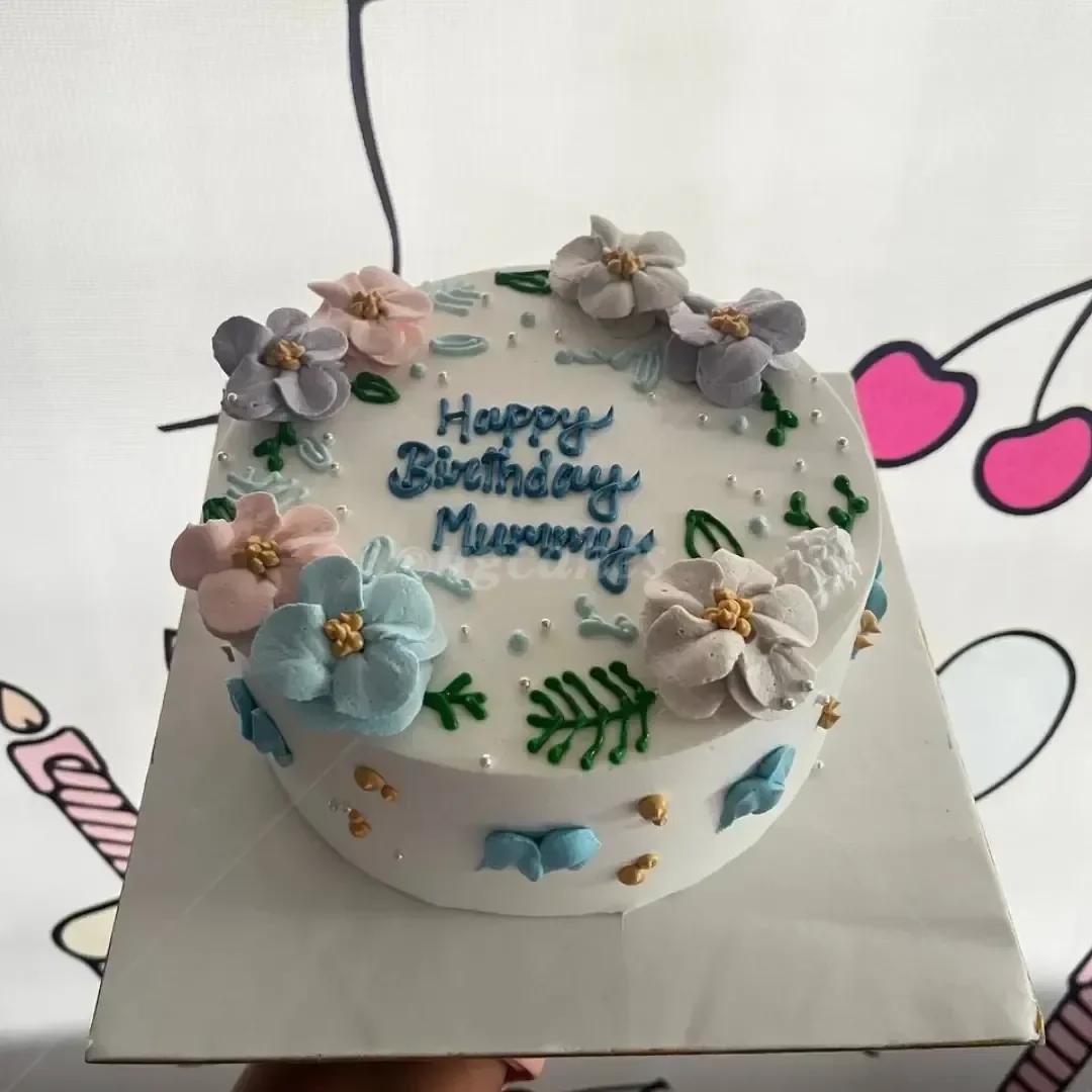 White minimalist birthday cake with pastel blue, pink, and lavender icing flowers and green leaves, featuring a blue piped message.