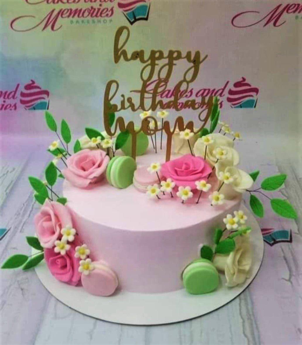 Pink floral birthday cake for Mom with pink and white gumpaste roses, green macarons, and a gold Happy Birthday Mom topper.