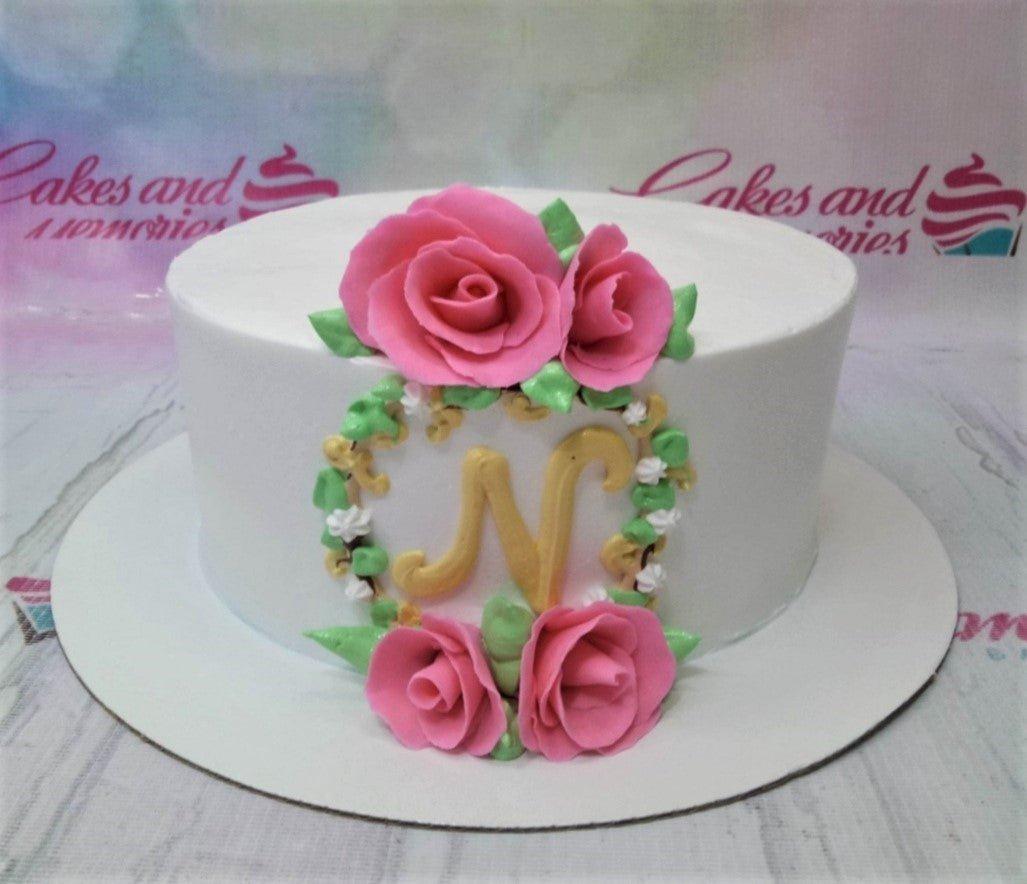 White minimalist cake with pink gumpaste roses and a gold letter N monogram surrounded by a floral wreath.