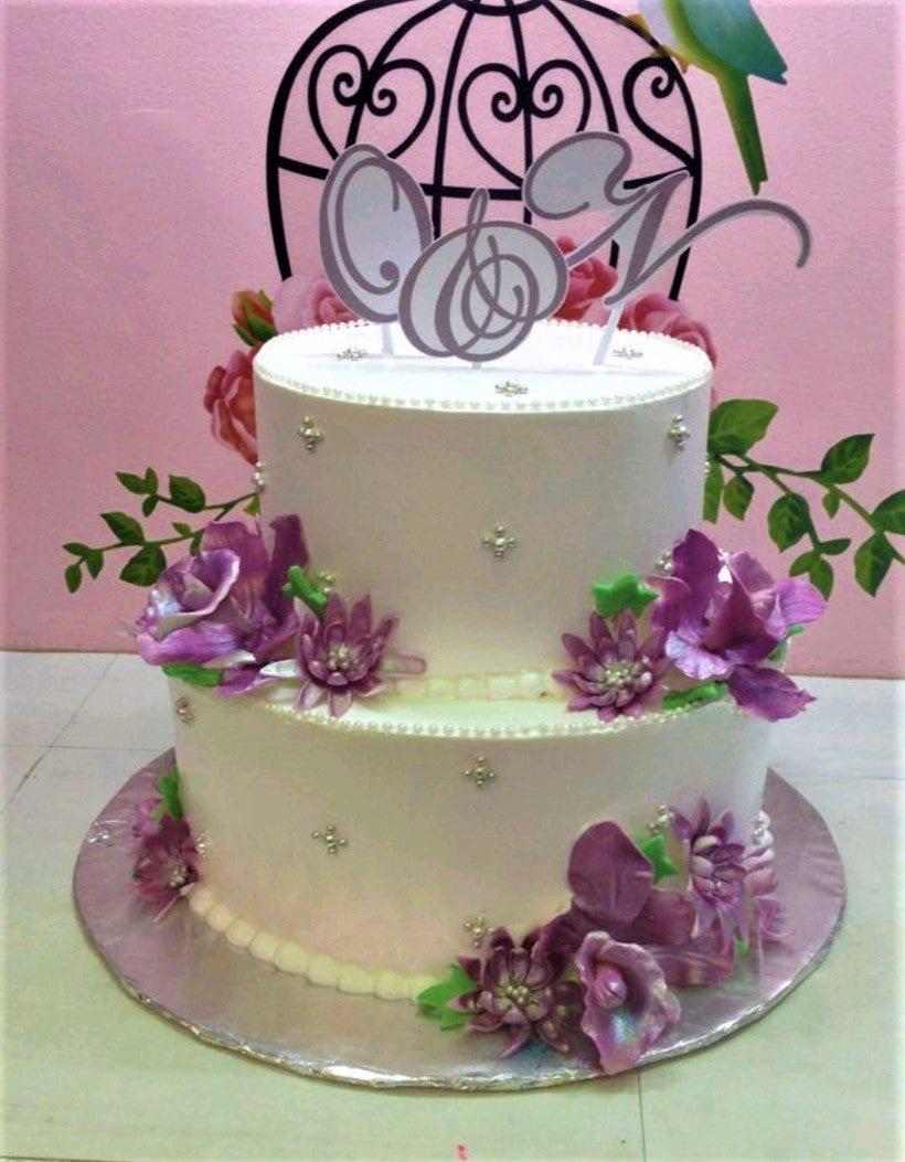 Elegant 2-tier white floral cake with purple gumpaste flowers, silver dragees, and a custom monogram topper.