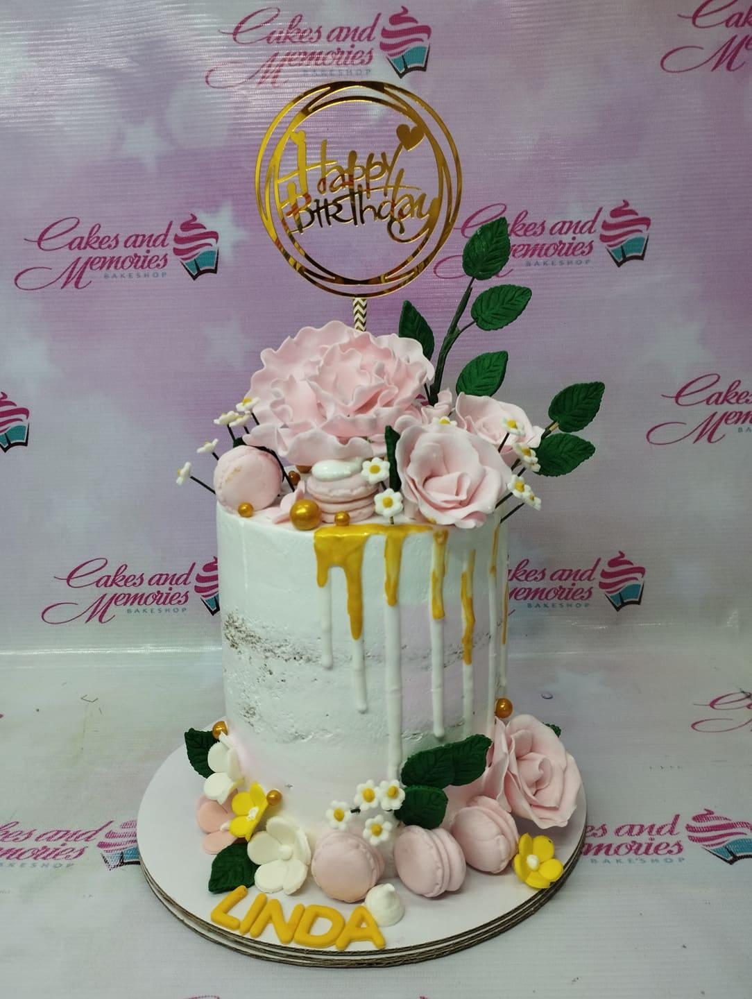 Tall white semi-naked cake with pink gumpaste roses, macarons, gold drips, and a gold Happy Birthday topper.