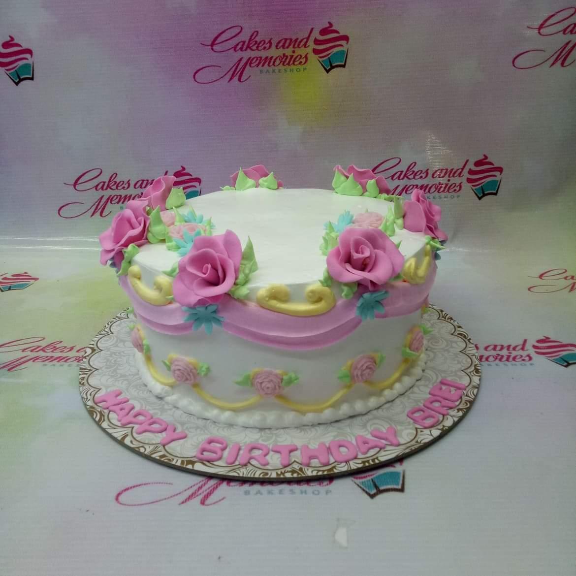 White round cake with pink icing roses, yellow swags, and a pink birthday message on the base board.