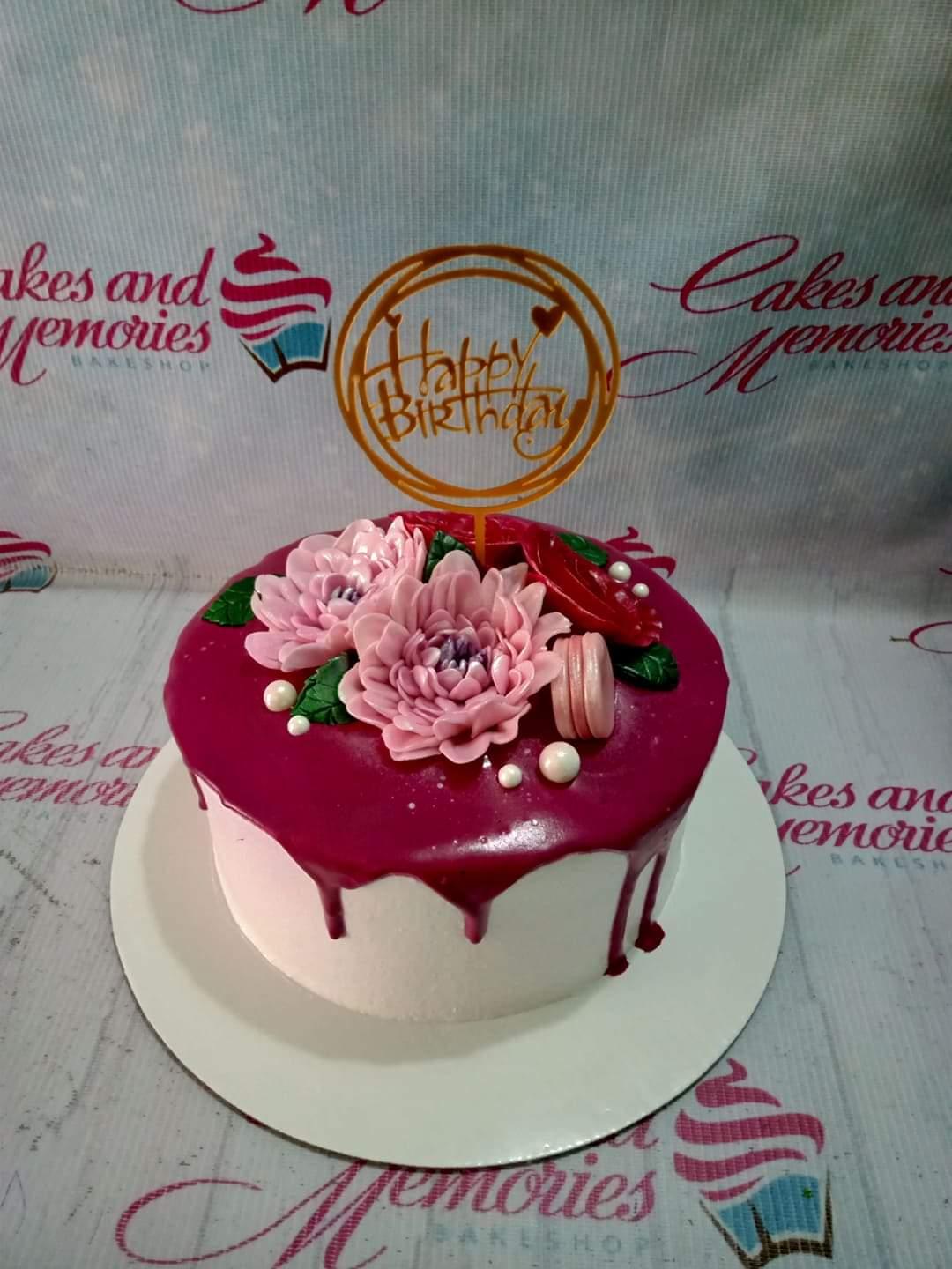 Single tier white cake with dark red drip, pink gumpaste flowers, a pink macaron, and a gold Happy Birthday topper.