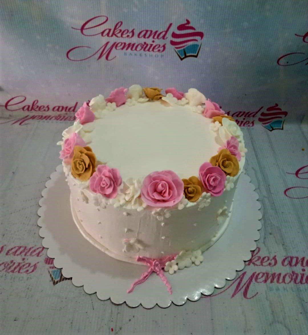 White round cake decorated with pink, gold, and white gumpaste roses and small white flowers around the top edge and base.
