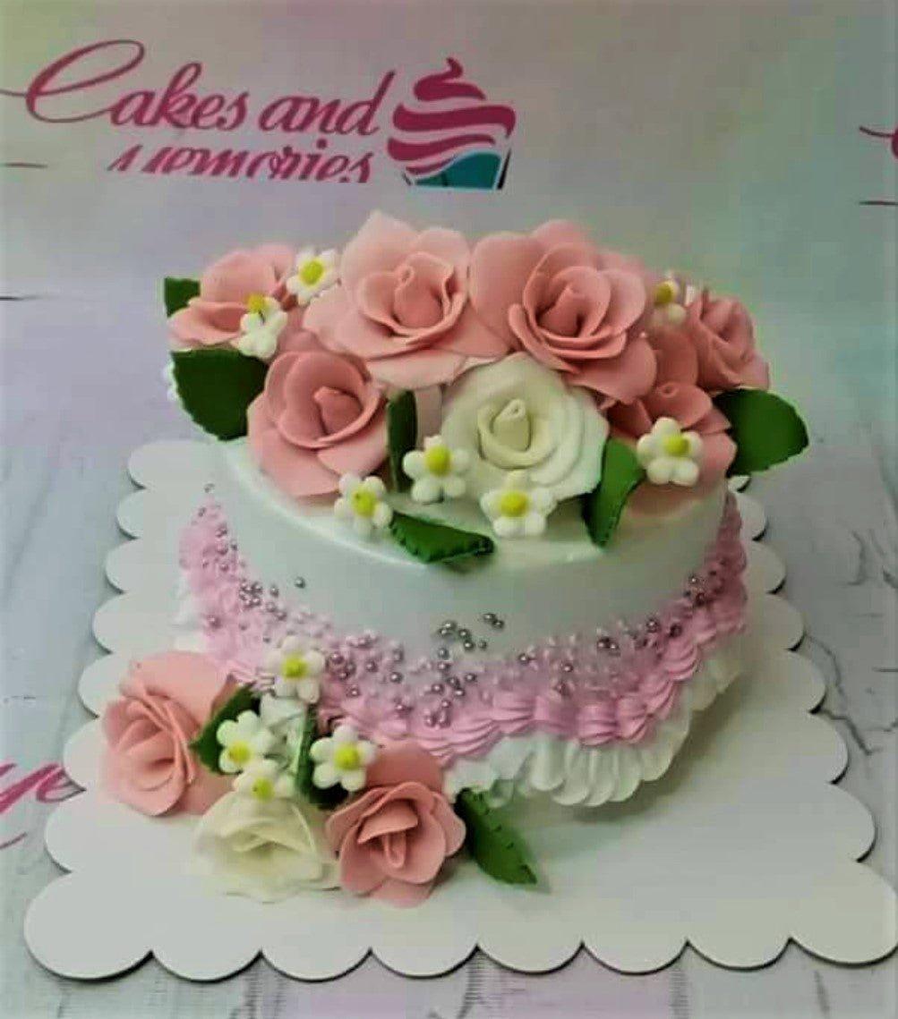 White round cake decorated with pink and white gumpaste roses, green leaves, and pink icing ruffles with silver dragees.