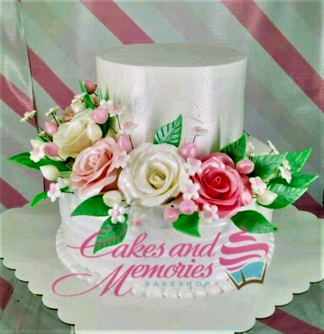 Elegant two-tier white cake decorated with large pink and white gumpaste roses and green leaves.
