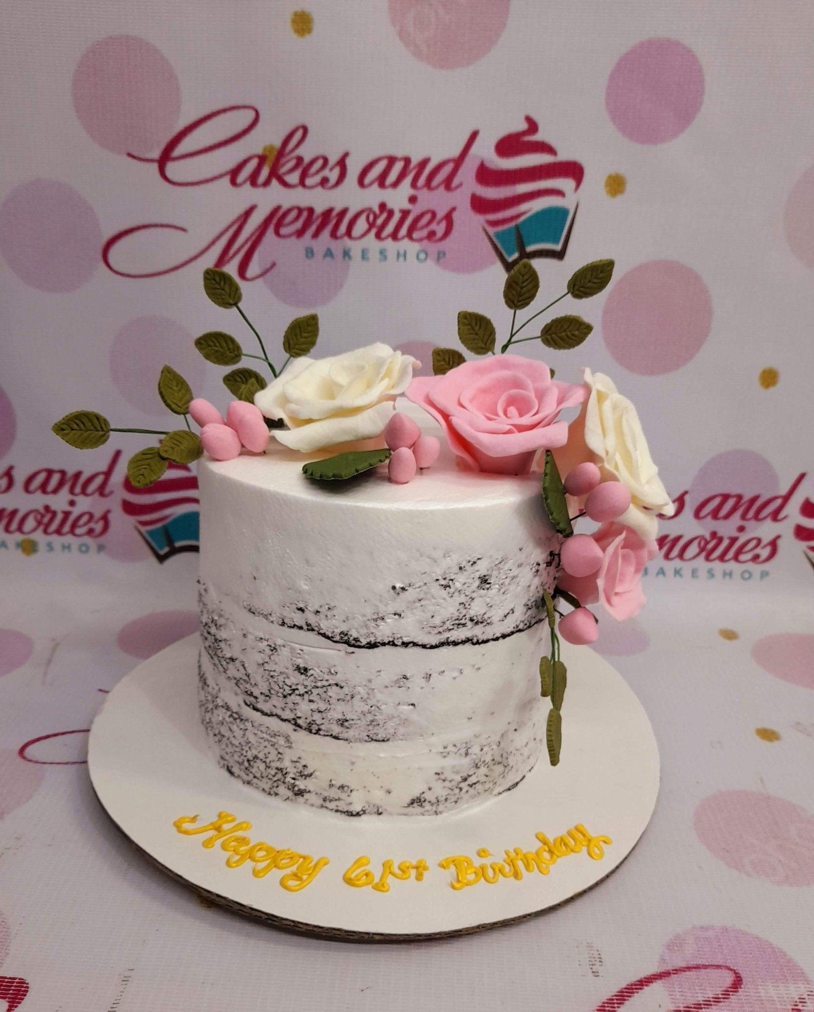 Semi-naked chocolate cake with white icing, pink and white gumpaste roses, green leaves, and a yellow birthday message on the base board.