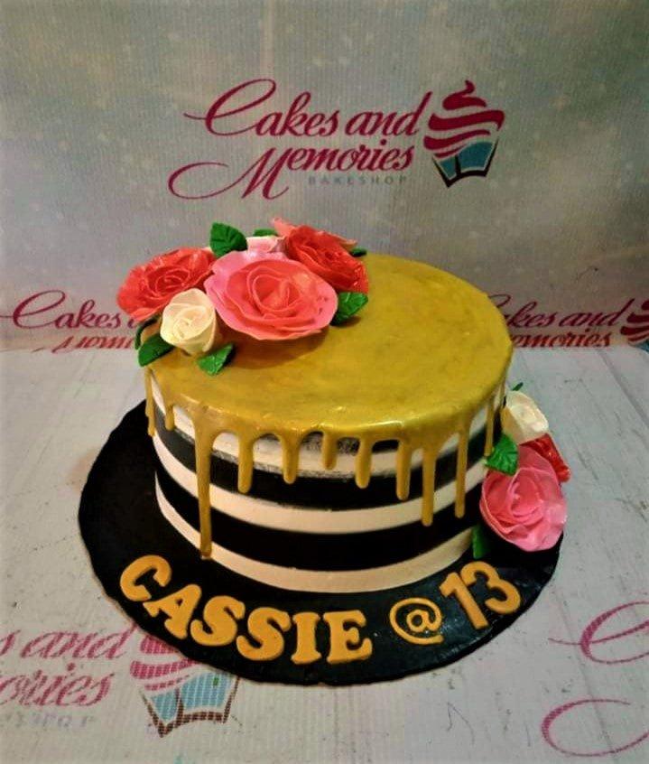 Black and white striped cake with gold drip, pink and white roses, and gold lettering on a black base board.