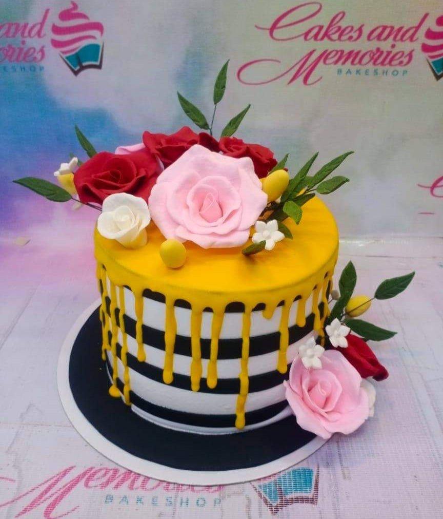 A single-tier cake with black and white horizontal stripes, yellow drip icing, and pink, red, and white gumpaste roses on top and side.