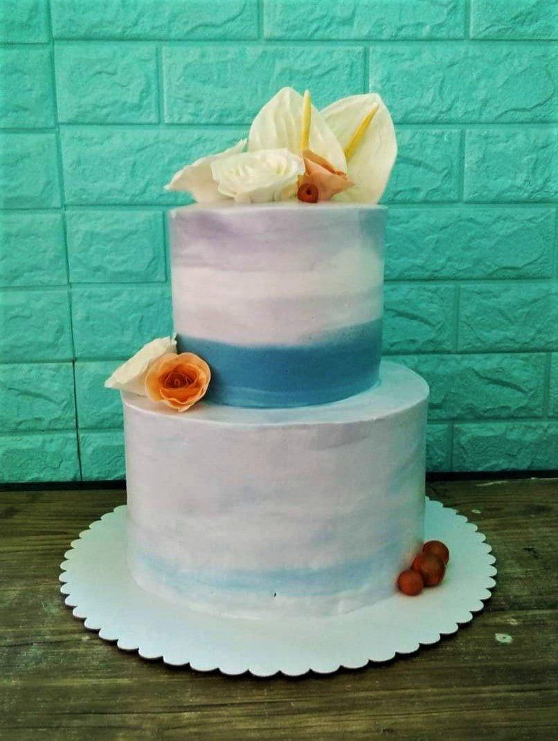 Two-tier watercolor cake with blue and purple ombre icing, topped with white and peach edible flowers and berries.