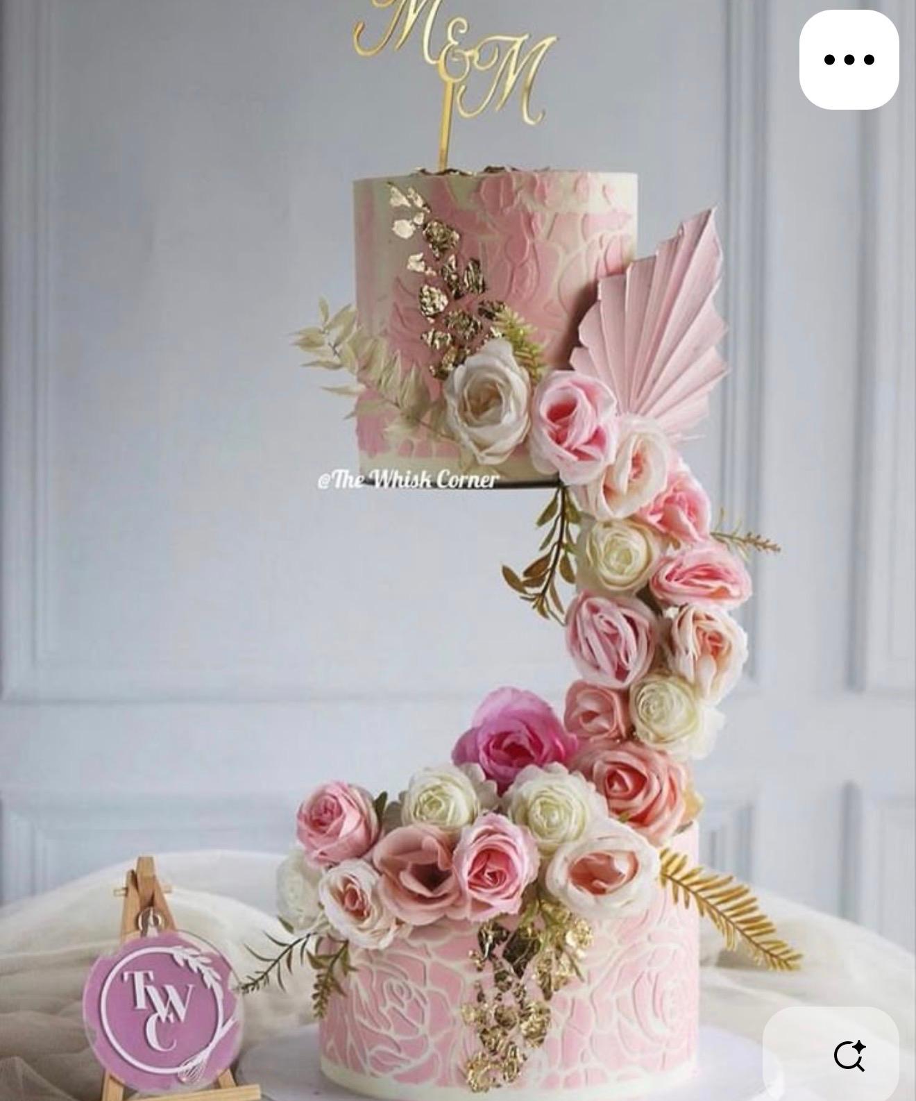Elegant two-tier pink and white cake with intricate floral patterns, gold leaf accents, and a cascading arrangement of pink and white roses.