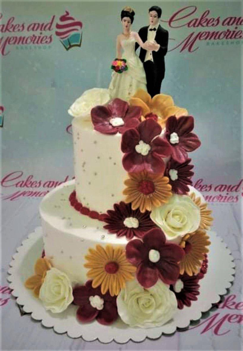 Elegant 2-tier white wedding cake decorated with burgundy, orange, and white gumpaste flowers and a bride and groom topper.