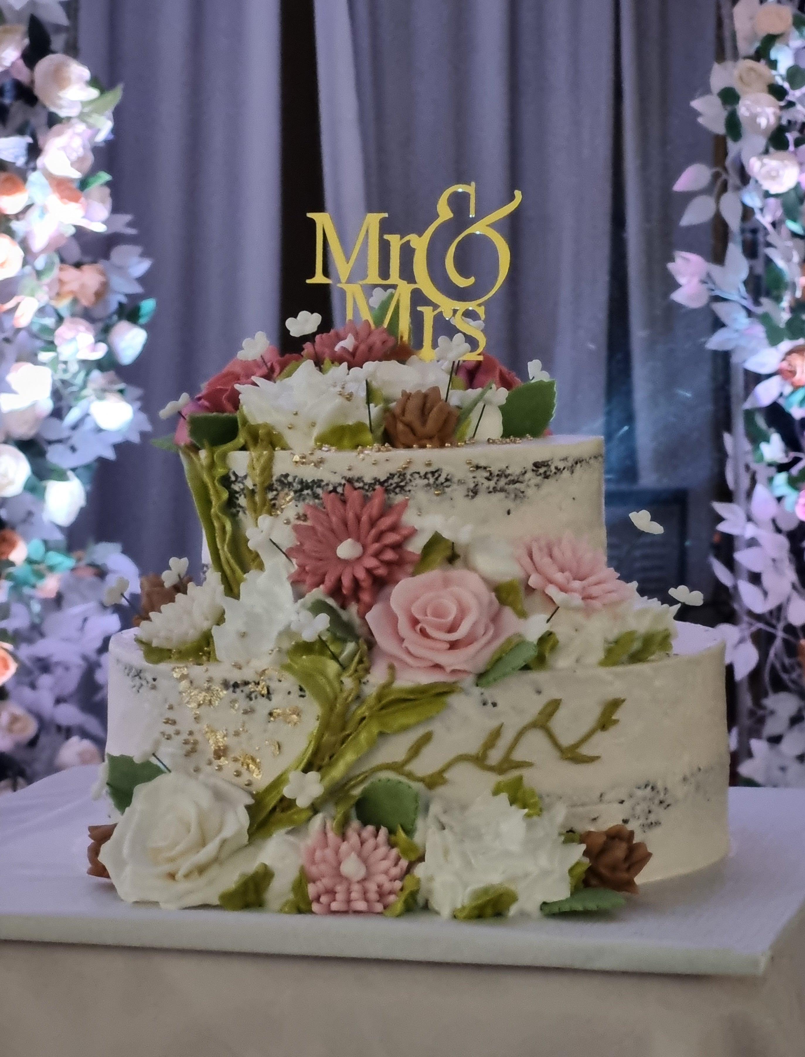 Elegant two-tier semi-naked wedding cake adorned with pink and white edible flowers, green leaves, and a gold Mr & Mrs topper.