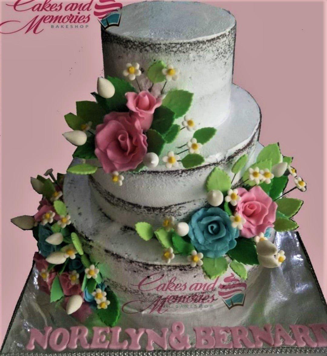 Three-tier semi-naked chocolate cake decorated with pink and teal gumpaste roses, white filler flowers, and green leaves.