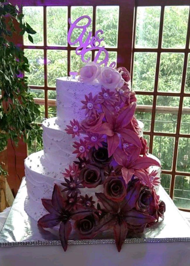 Elegant 3-tier white wedding cake with a cascading floral arrangement of purple and burgundy gumpaste flowers and a purple script topper.