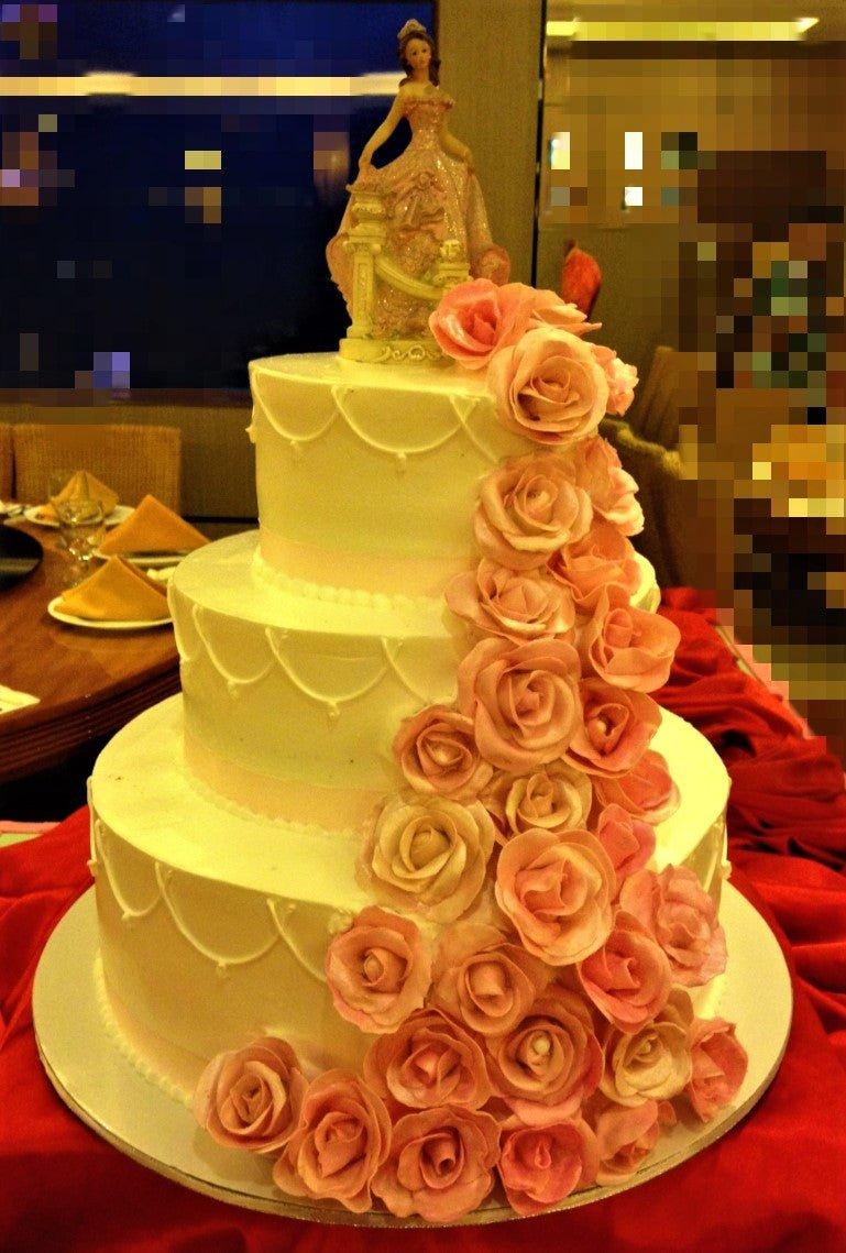 Elegant three-tier white wedding cake adorned with a cascading trail of peach and pink edible roses and a princess figurine topper.